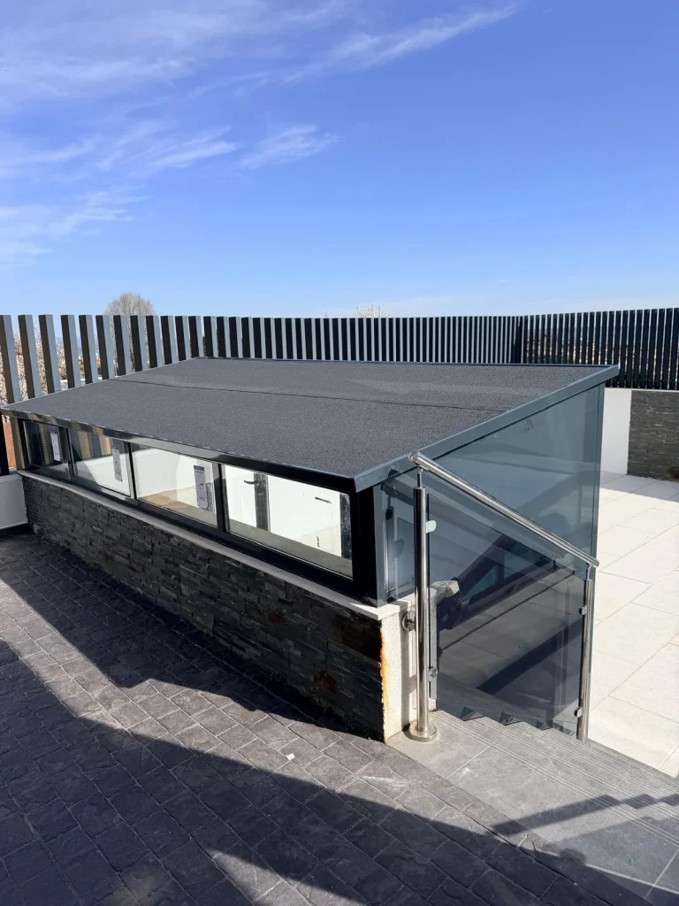 Cerramiento de aluminio y vidrio con techo inclinado sobre un murete de piedra que cubre la bajada a un sótano