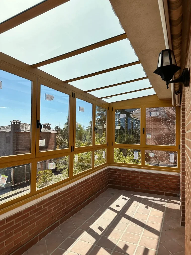 Interior de una terraza cerrada con perfiles de aluminio color madera, ventanas correderas y techo de vidrio