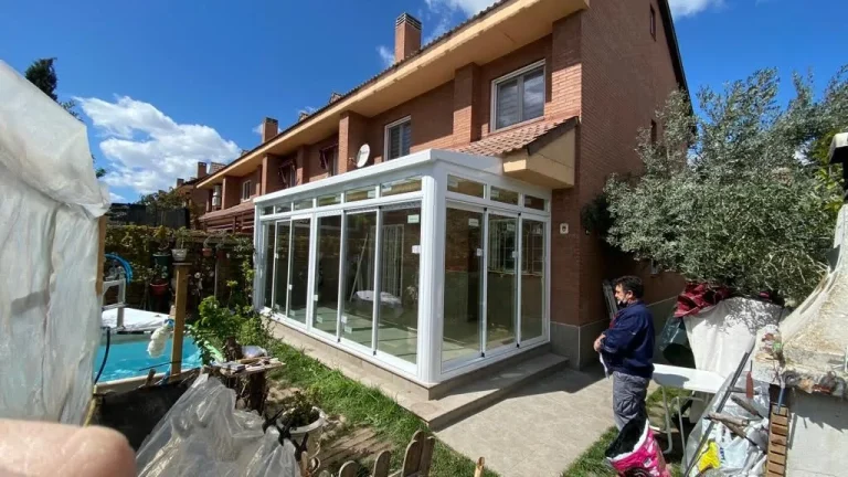 Porche de una casa adosada cerrado con aluminio blanco y techo acristalado, visto desde el jardín