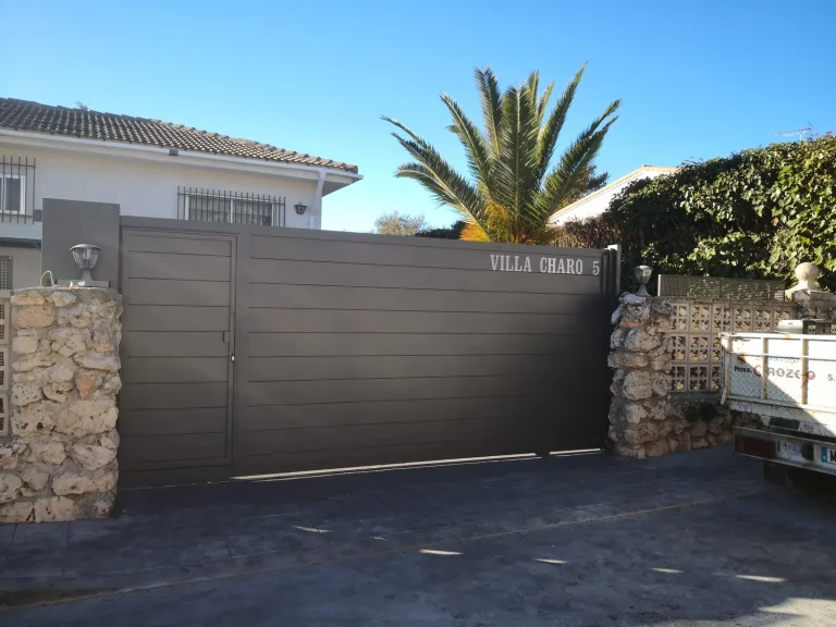Puerta de vehículos de aluminio soldado gris oscuro con puerta peatonal integrada, entre muros de piedra