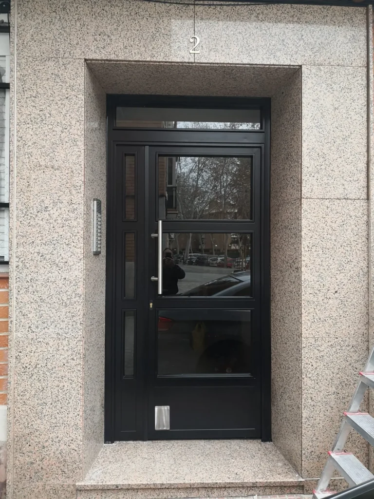 Puerta de portal de aluminio negro con vidrio y tirador vertical, enmarcada en granito
